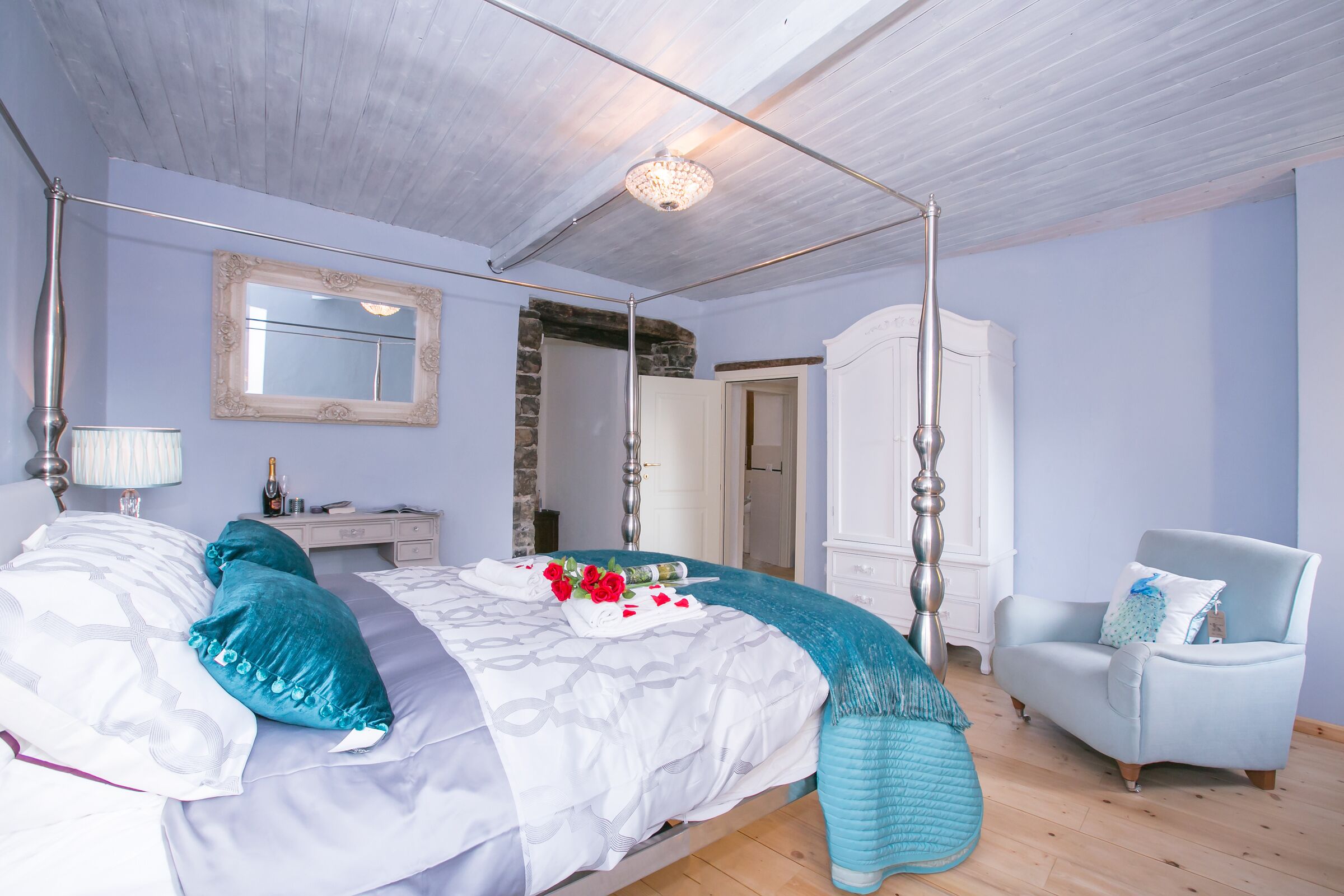 A four-poster bed with a canopy, a white dresser, a light blue armchair, and a stone wall.