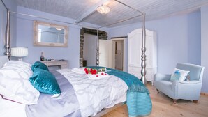 Una cama con dosel, un aparador blanco, un sillón azul claro y una pared de piedra.