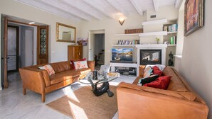 Una sala de estar con un sofá de cuero marrón, una mesa de centro de vidrio y una chimenea. Hay estanterías y un televisor montado en la pared.