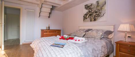 A bedroom with a large bed, a chandelier, a radiator, a wooden dresser, and a painting on the wall.