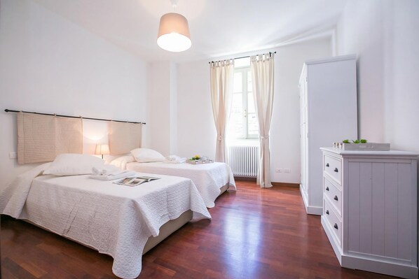 A bedroom with two beds, a bedside table with a lamp, a dresser, and a window with curtains. - Casa Al Prato Apt. B (Varenna)