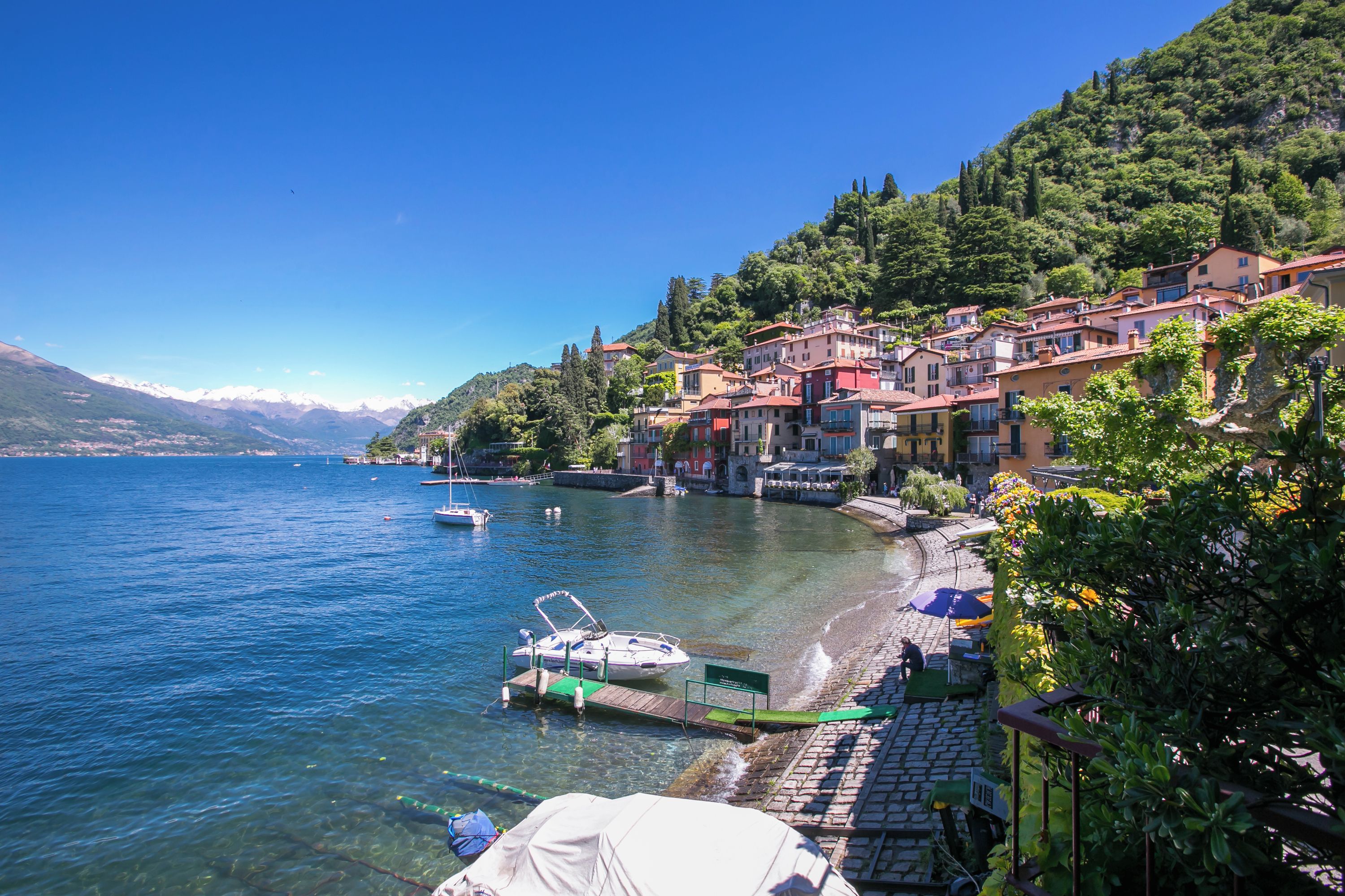 Un pueblo en la orilla de un lago, con edificios a lo largo de la costa, barcos amarrados y un cielo azul despejado.