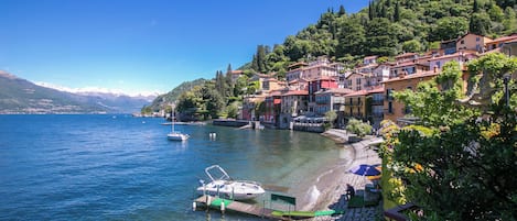 Un pueblo en la orilla de un lago, con edificios a lo largo de la costa, barcos amarrados y un cielo azul despejado.