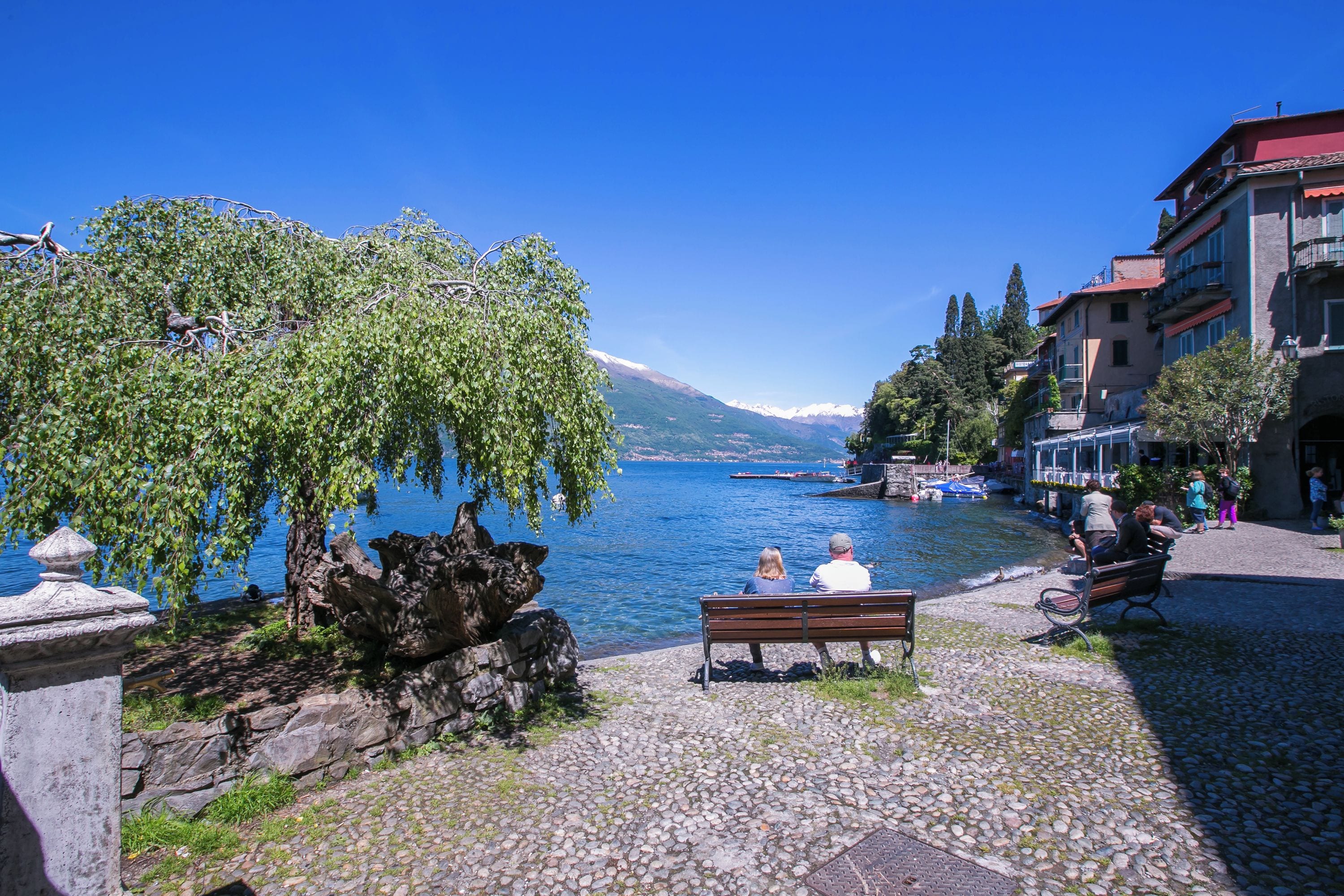 Una passeggiata sul lungolago lastricata, con panchine, una balaustra in pietra e edifici con balconi che si affacciano sul lago.