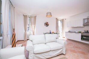 A living room with a white sofa, a dining table with chairs, a kitchen area with cabinets and appliances, and a window with curtains. - Casa Al Prato Apt. G (Varenna)
