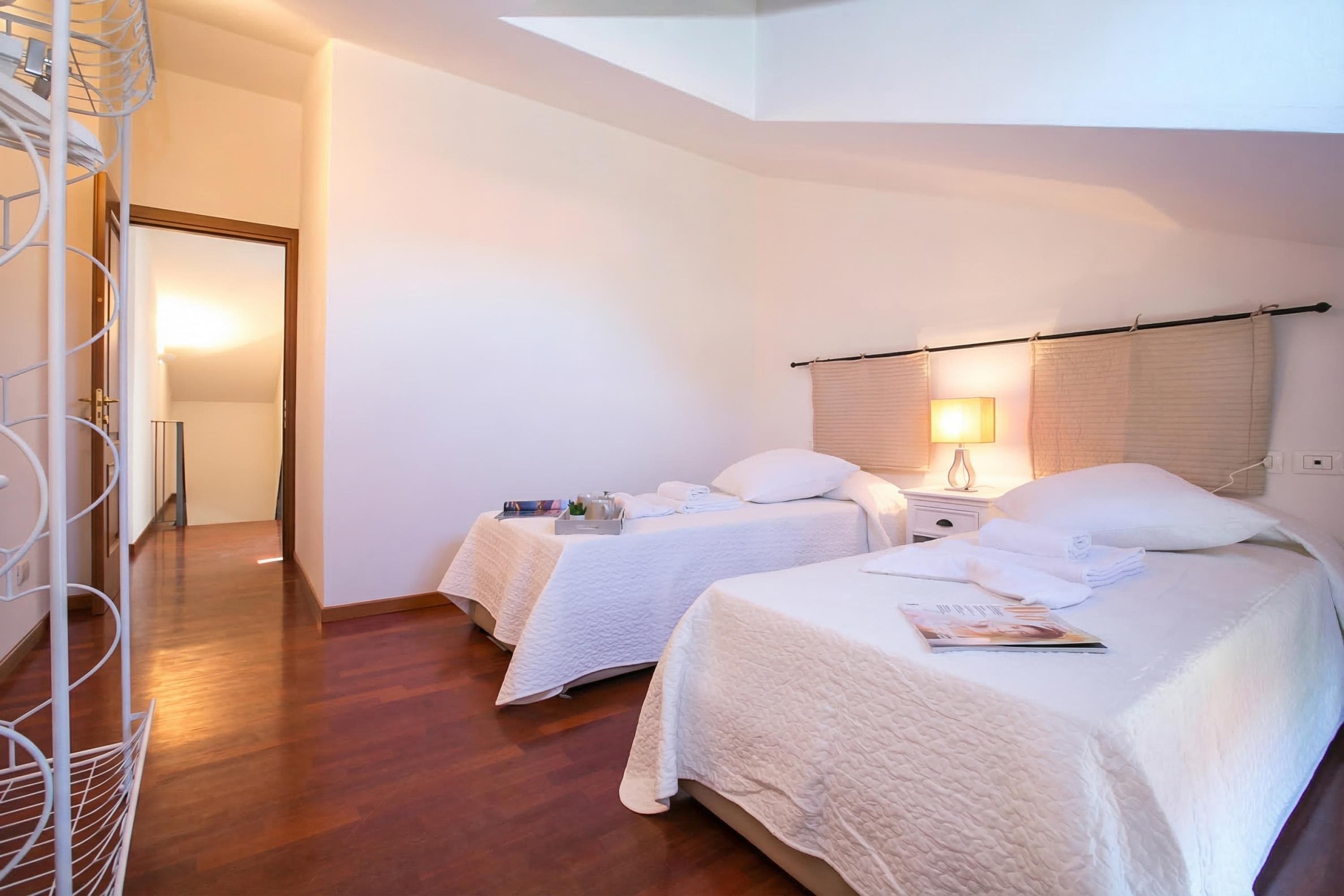 Habitación de hotel con dos camas, piso de madera, pared blanca, una puerta y una mesita de noche con lámpara.