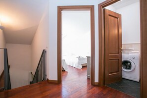Un pasillo con una escalera de madera, un cuarto de lavado con lavadora y una cama al fondo.