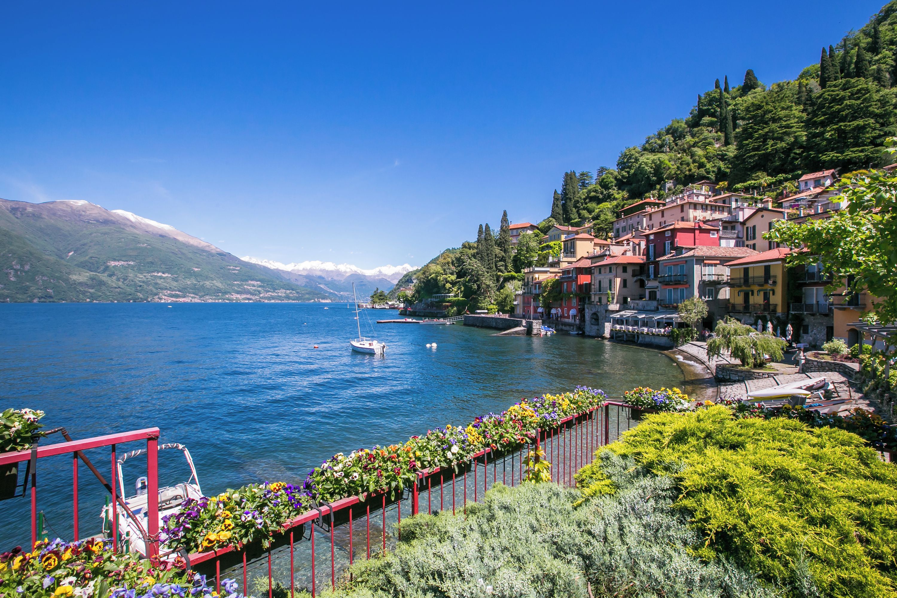 A lakeside town with buildings along the shore, a dock with a boat, and a row of flower-lined railings.