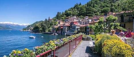Un pueblo a orillas de un lago, con edificios a lo largo de la orilla, una barandilla roja adornada con flores y un cielo azul despejado.