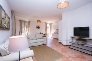 Un saló amb un sofà, una làmpada, un televisor sobre un suport, una cuina al fons i una zona de menjador.
