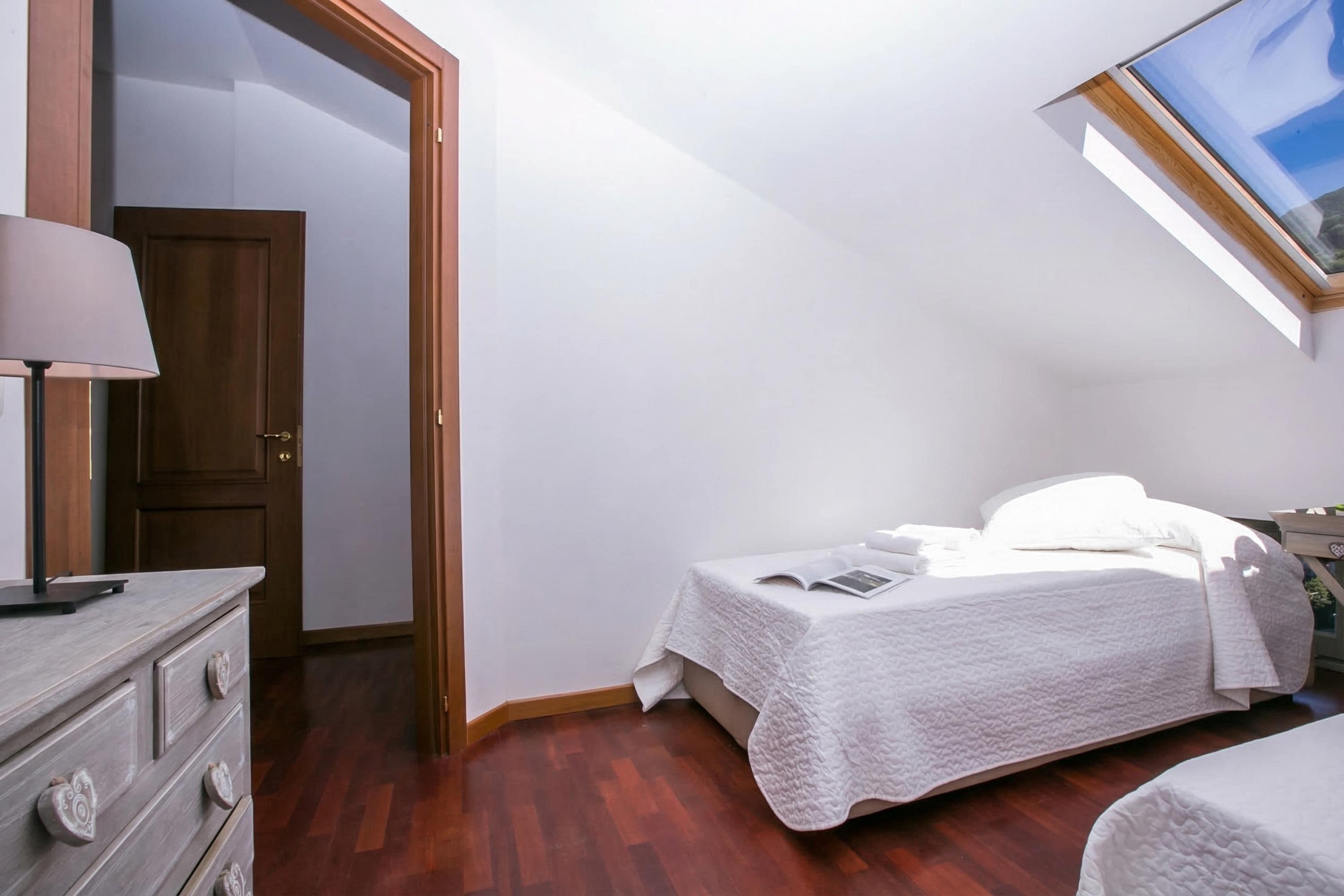 A bedroom with a wooden dresser, a bed with a white quilt, and a skylight.