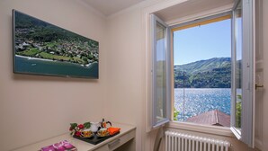 A room with a large window offering a view of a lake and mountains, a desk with a magazine, and a radiator. - Laglio Deluxe (Carate Urio)