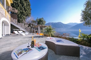 A terrace with a table set for two, a bottle of champagne, and magazines. There are lounge chairs and a pergola with a view of the sea and mountains. - Villa Lugano Lakefront  (Valsolda)