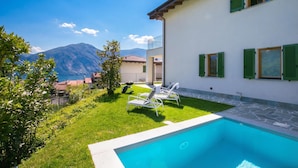 A poolside view with a clear sky, a mountain in the background, and a white building with green shutters. - Villetta Tara (Tremezzina)