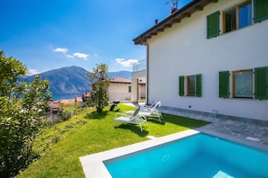 Vista a la piscina con cielo despejado, una montaña al fondo y un edificio blanco con contraventanas verdes.