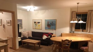 A living room with a dark-colored sofa, a wooden coffee table, and a dining table with chairs. There are two framed artworks on the wall, a ceiling light fixture, and a window with curtains.