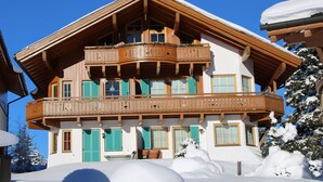 A chalet with a wooden balcony, multiple windows with shutters, and snow-covered surroundings.