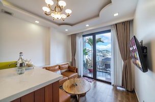 A modern hotel room with a dining area, a sofa, a TV mounted on the wall, and a balcony with a view.
