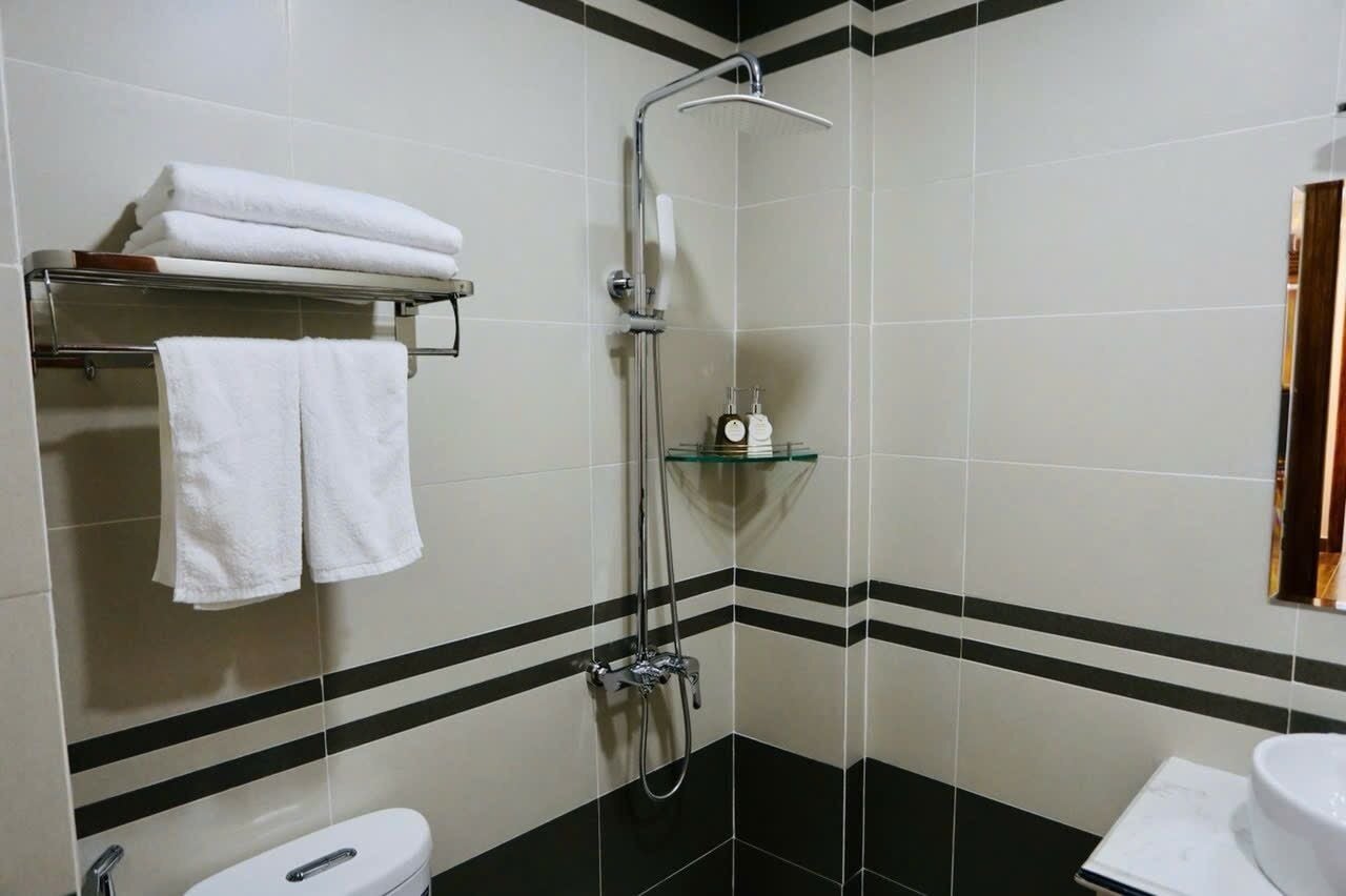 Un baño con ducha, dos lavamanos y un inodoro. Hay toallas en un perchero y un estante de vidrio con artículos de aseo.