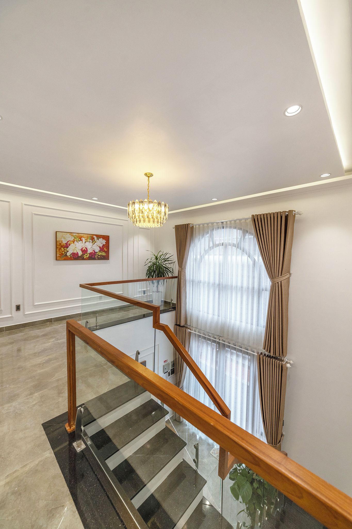 A modern staircase with glass railings and a wooden handrail, leading to a room with a large window and a chandelier.