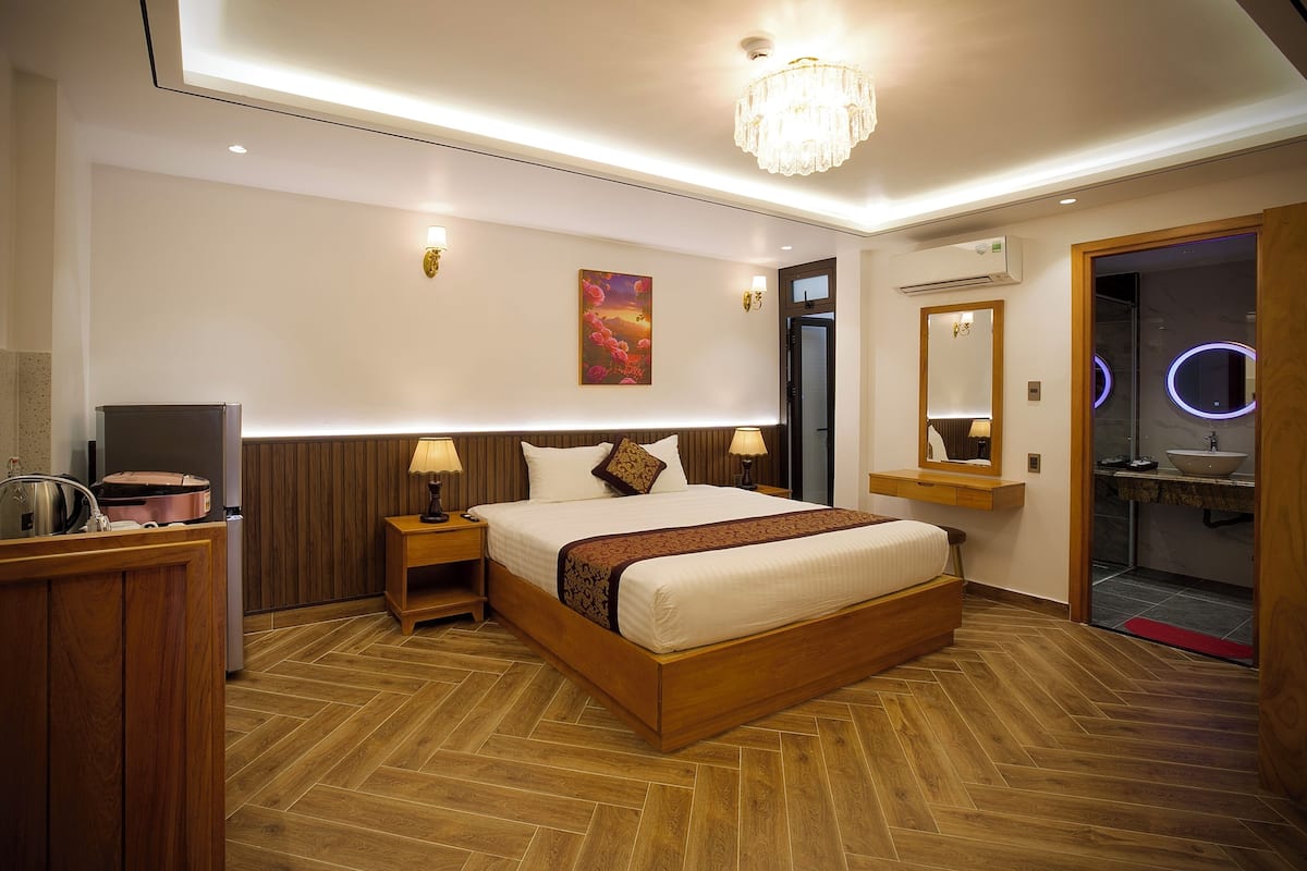 A modern hotel room with a wooden bed, bedside lamps, a chandelier, a painting on the wall, and a bathroom with a mirror and sink.
