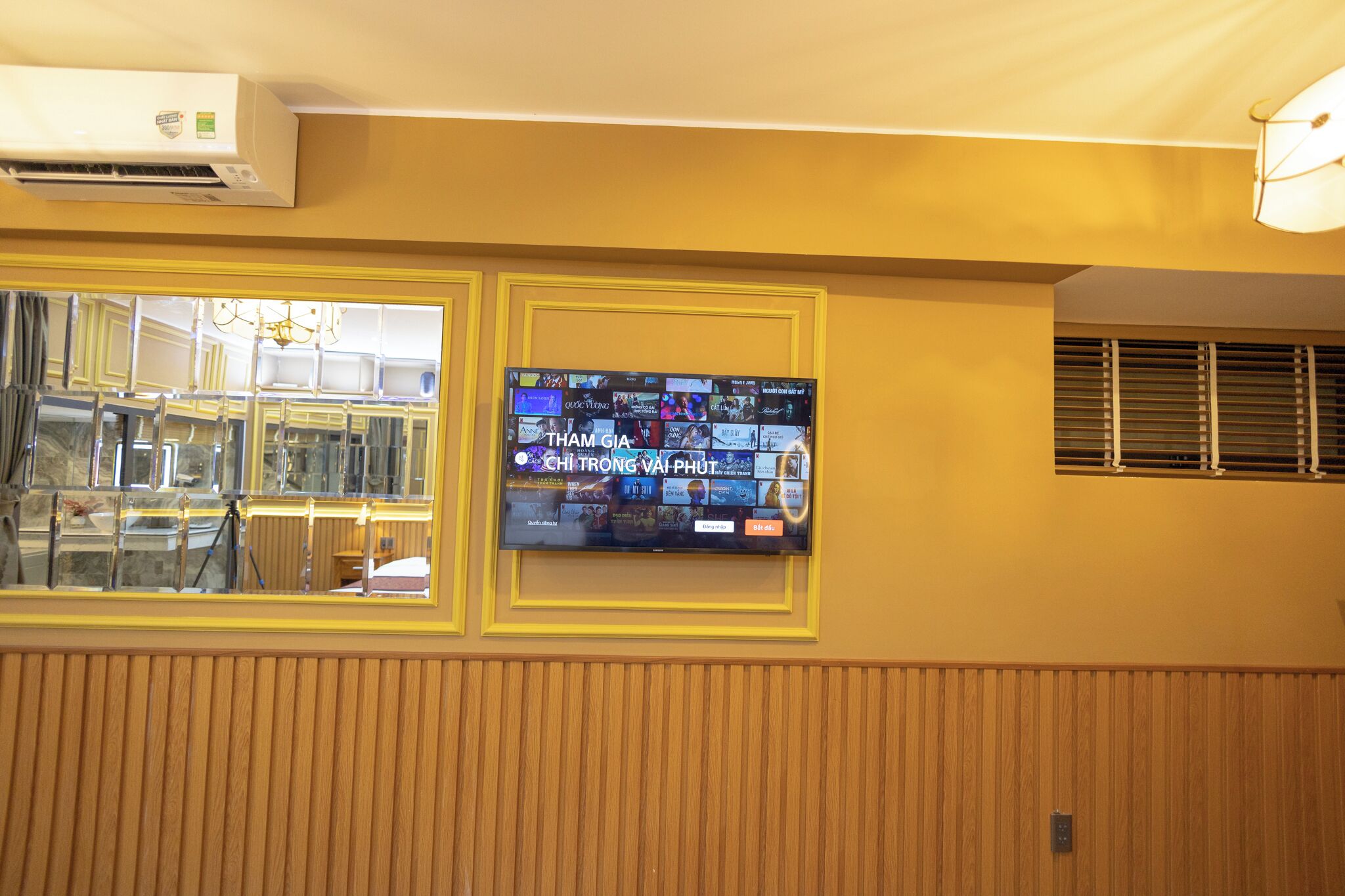 A wall-mounted television displaying a news channel, a window with blinds, and an air conditioning unit.