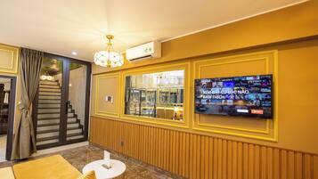 Un vestíbulo de hotel con una gran pantalla de televisión mostrando un canal deportivo, una escalera que conduce a otro piso y un sofá amarillo.