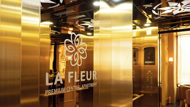 Interior de un ascensor con una puerta de vidrio que muestra “LA FLEUR PREMIUM CENTRAL APARTMENT” y un logotipo floral.
