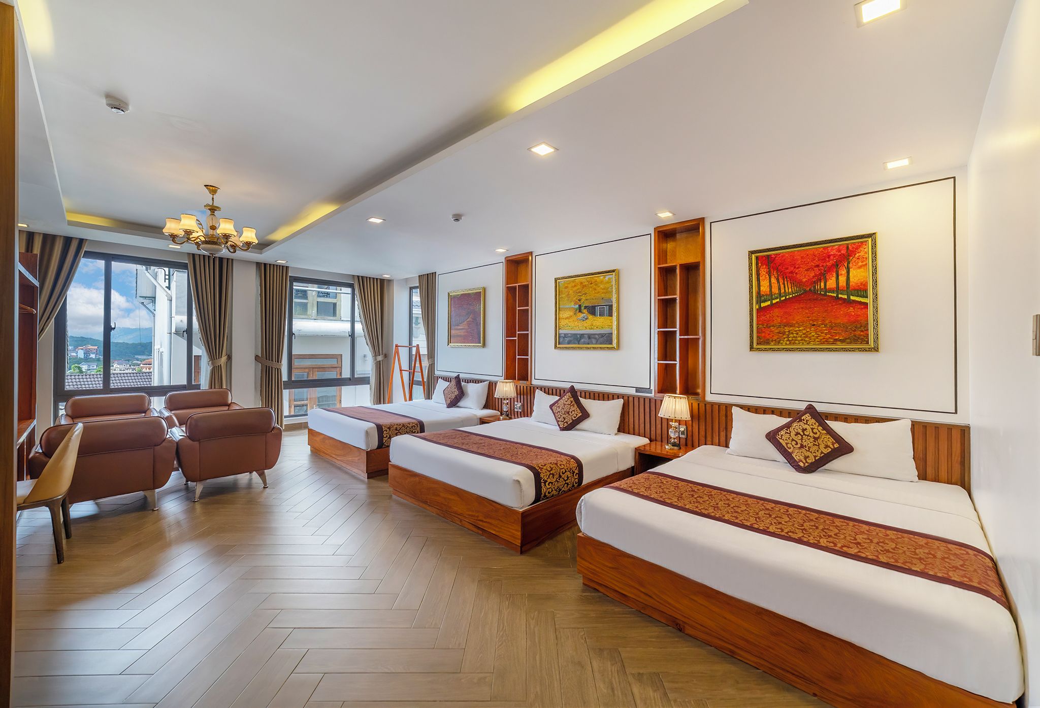 A hotel room with a large bed, a smaller bed, a sofa, and a desk. There are paintings on the walls and a view of the city through the windows.