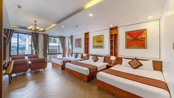 A hotel room with a large bed, a smaller bed, a sofa, and a desk. There are paintings on the walls and a view of the city through the windows.
