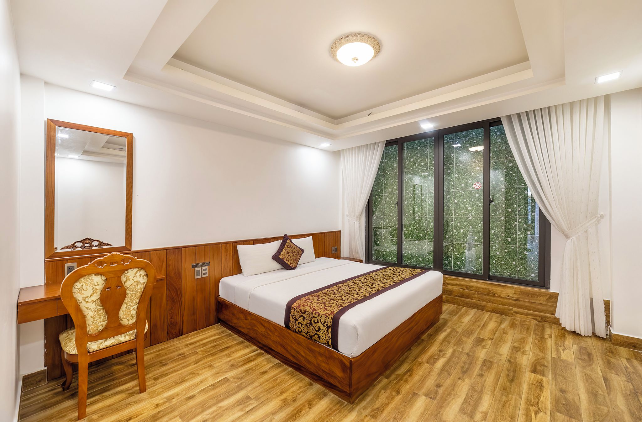 Habitación de hotel con una cama de madera, un escritorio, una silla y un gran ventanal con cortinas.