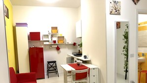 Une cuisine compacte avec des meubles blancs, un réfrigérateur rouge et un petit coin repas comprenant une table blanche et une chaise rouge.