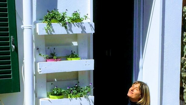 Une personne assise sur un banc devant un bâtiment orné de plusieurs étagères à plantes.