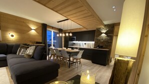 Ein modernes Wohnzimmer mit einem dunklen Sofa, einem Glastisch mit Kerzen und einem Essbereich mit Küche im Hintergrund.