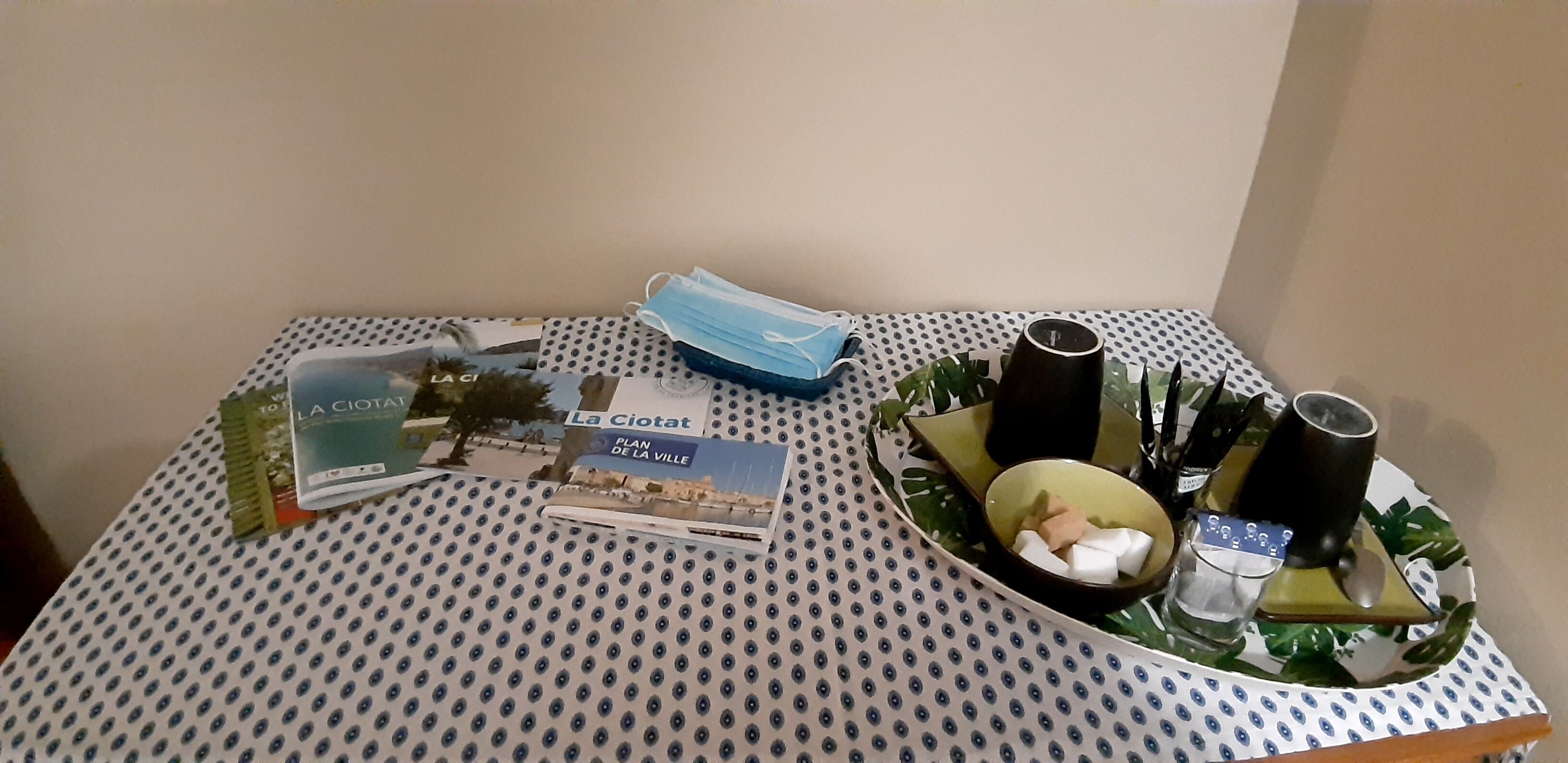 Une table recouverte d’une nappe à pois, une brochure intitulée « LA CIOTAT », un masque facial bleu et un plateau avec une tasse, des morceaux de sucre et un verre.
