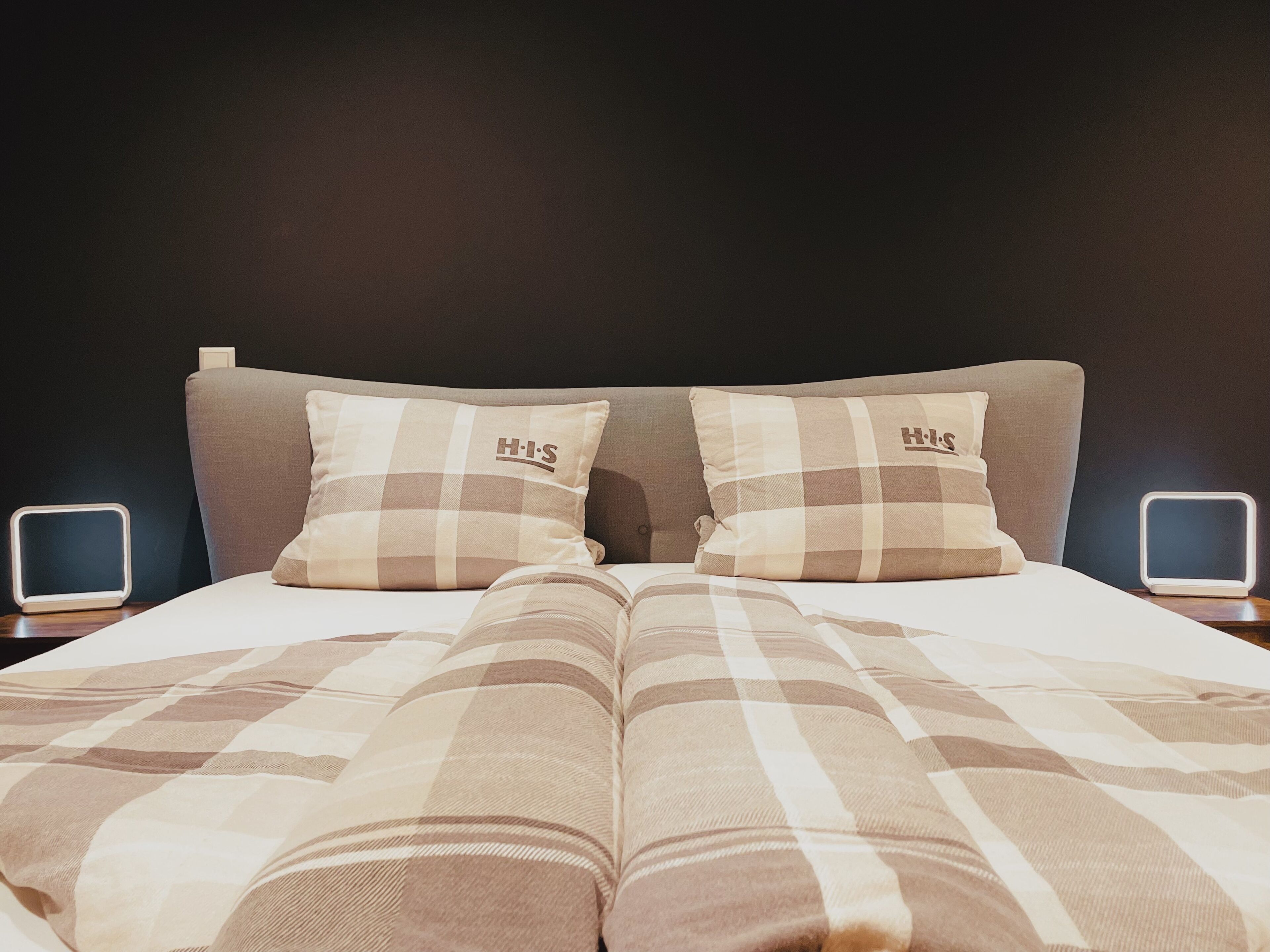 Un letto con una coperta a quadretti e due cuscini con un logo, affiancato da comodini con schermi illuminati.