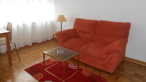 Een woonkamer met een rode bank, een glazen salontafel, een houten stoel, een lamp en een schilderij aan de muur.