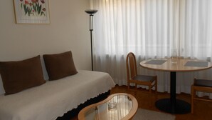 Een slaapkamer met een bed, twee bruine kussens, een nachtkastje met een glas, een ronde tafel met twee stoelen, een vloerlamp en een raam met dunne gordijnen.
