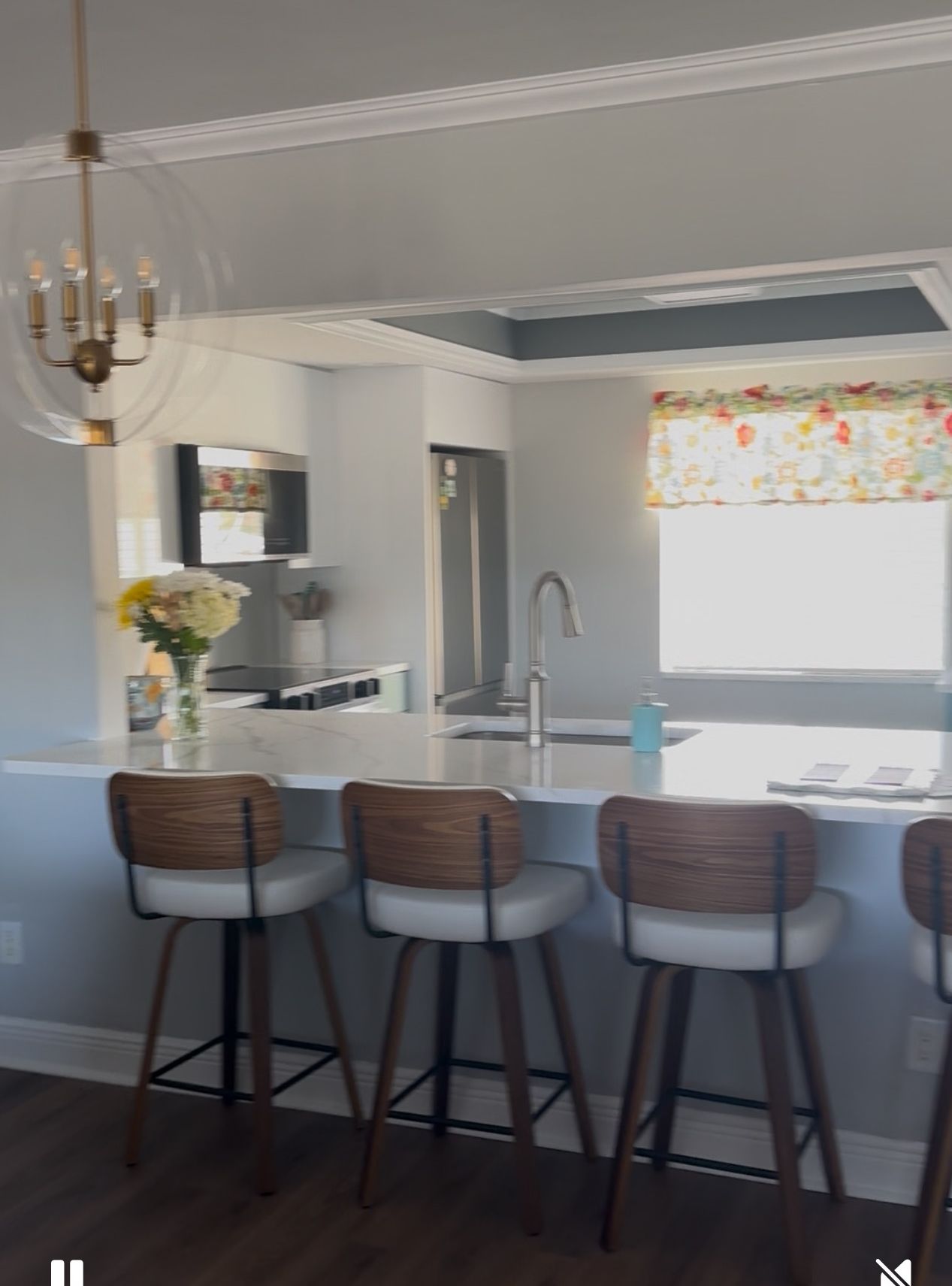 A modern kitchen with a central island, bar stools, a chandelier, and a window with a floral curtain.