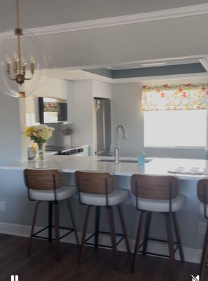 A modern kitchen with a central island, bar stools, a chandelier, and a window with a floral curtain.