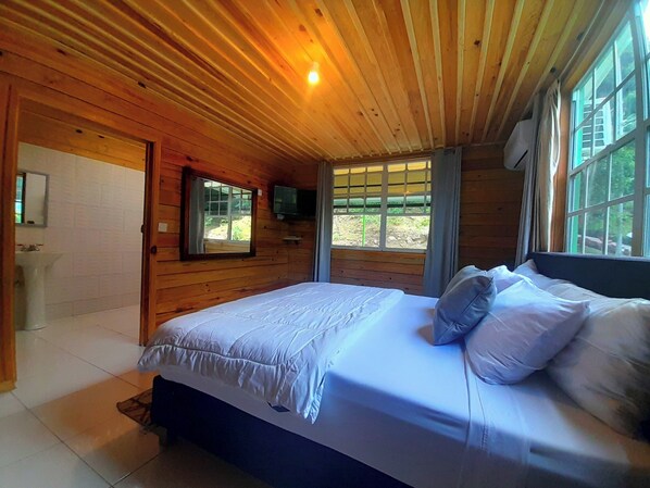 A wooden cabin bedroom with a bed, a television, and a window. - Liberty Suite Cottage 8 (Roseau)
