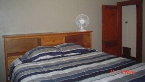 A bedroom with a wooden bed frame, a striped bedspread, and a standing fan on a nightstand.