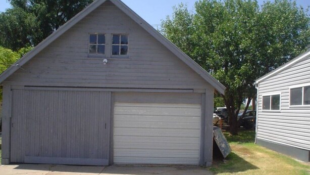 A single-story garage with a large garage door and a smaller door, surrounded by a concrete driveway and trees.