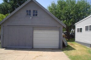 A single-story garage with a large garage door and a smaller door, surrounded by a concrete driveway and trees.
