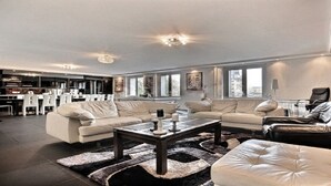 Ein großzügiges Wohnzimmer mit einer großen Sofagarnitur, einem Couchtisch und einem Ledersessel. Im Hintergrund ist eine Küchenzeile mit modernen Geräten und ein Essbereich mit weißen Stühlen zu sehen.