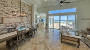 A spacious living room with a large window overlooking a beach, a dining area with a brick wall, and a ceiling fan.