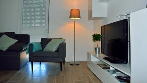 Una sala de estar moderna con un sofá, una lámpara de pie, un televisor sobre un soporte blanco y una ventana con persianas.