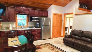 Cocina compacta con armarios de madera oscura, refrigerador de acero inoxidable y microondas. Hay una mesa de comedor con sillas y un sofá de cuero marrón en la sala de estar.