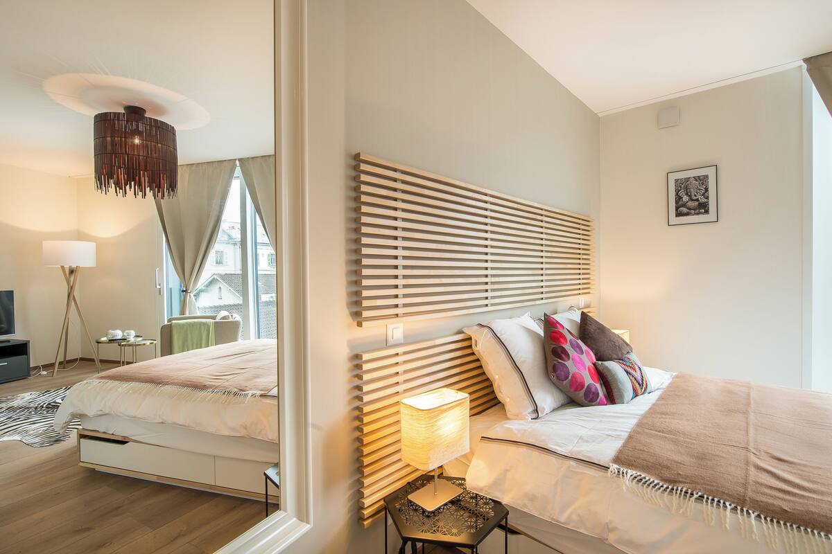 A modern bedroom with a large bed, a bedside table with a lamp, and a view of the outside through large windows.