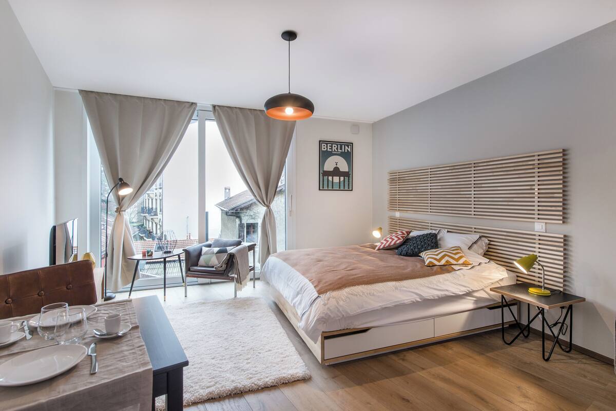 A modern bedroom with a large bed, a dining area with a table set, and a view of the city through large windows.